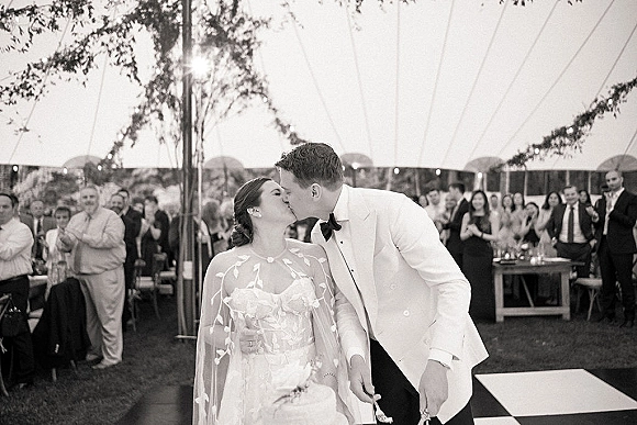 Wedding cake cutting as bride in embroidered cape and groom in white tux slice a tiered cake under string lights in a tent reception