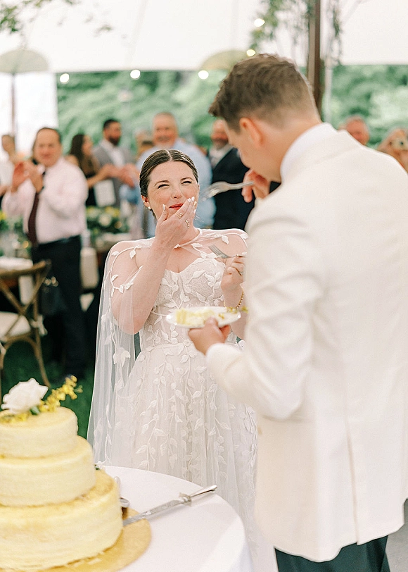 Cake cutting moment as bride in lace dress with cape veil and groom in white suit slice floral-topped cake under string lights in a tented reception