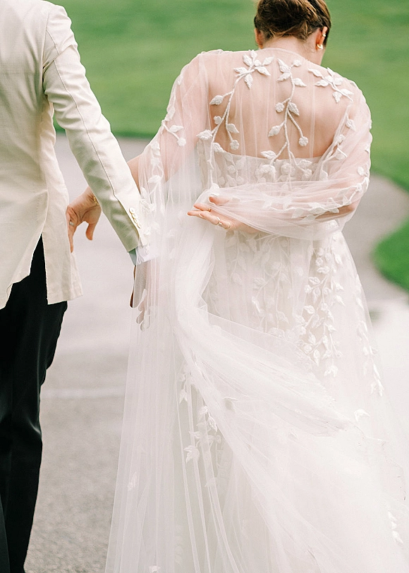 Couple walking away, bride and groom walking hand in hand on an outdoor path, tulle veil trailing over floral applique dress and light suit jacket