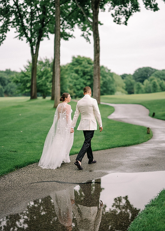 Couple portrait of bride and groom walking hand in hand, her veil trailing over a tree-lined path with puddle reflections on an overcast day
