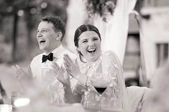 Reception candid moment of the bride and groom laughing at a toast, bride in veil and dress, groom in bow tie amid soft drapery lights