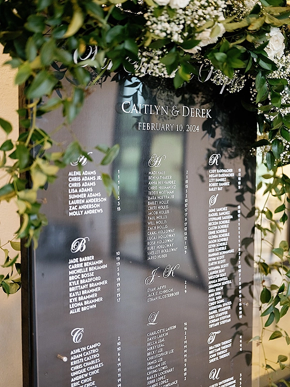 Wedding seating chart acrylic sign with calligraphy lettering, framed by white flowers and baby’s breath against a mirrored backdrop in soft indoor light