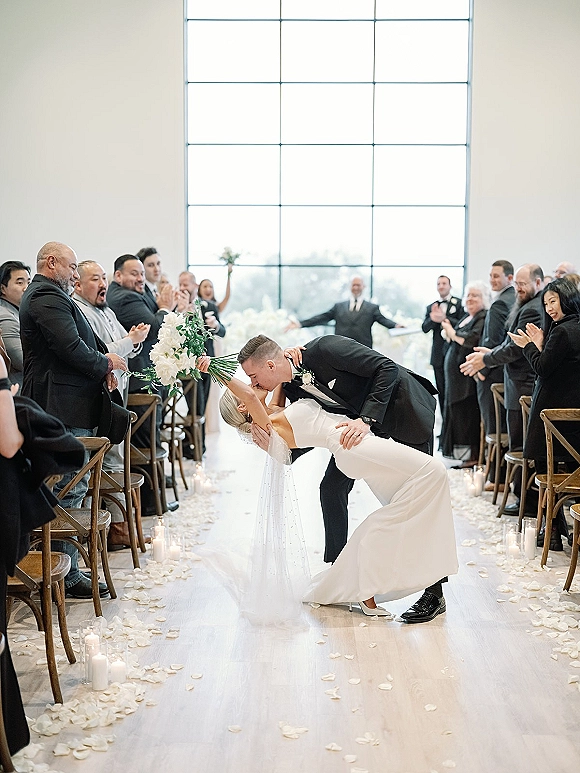 Wedding kiss portrait of groom dipping bride in a dip kiss wedding photo, bouquet raised, long veil flowing down a candlelit aisle beneath a grid window