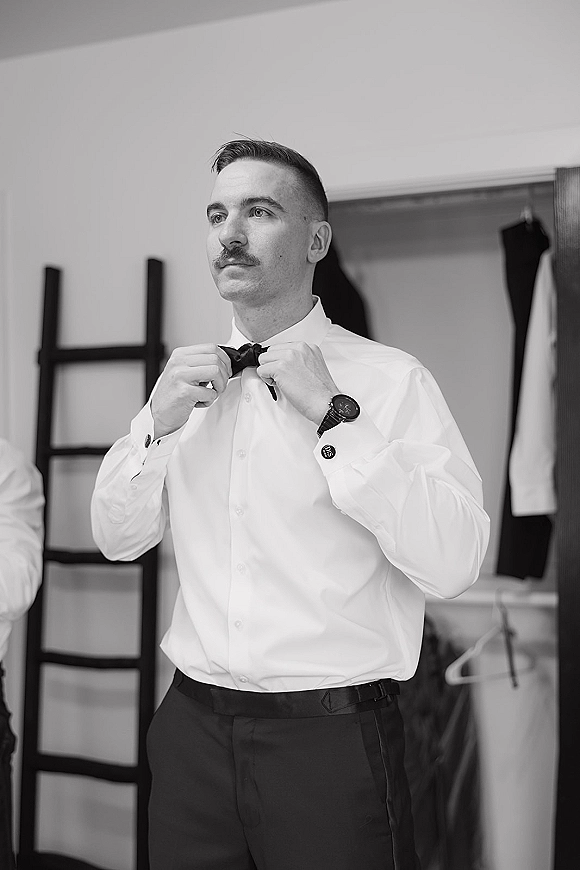 Groom getting ready, adjusting a bow tie on his dress shirt with a wristwatch visible, in a closet dressing room with hangers
