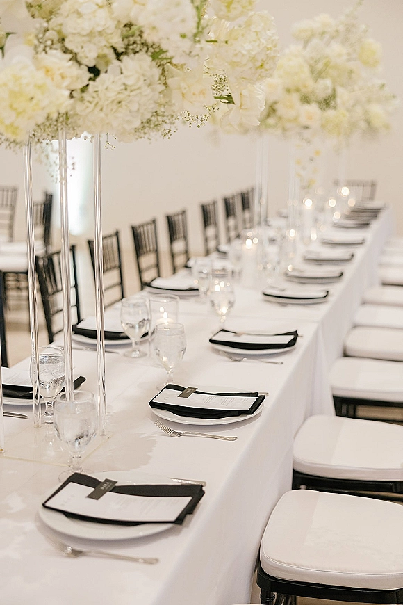 Reception tablescape with long banquet table setup, tall white floral centerpieces in clear vases, black napkins, candles, and glassware indoors