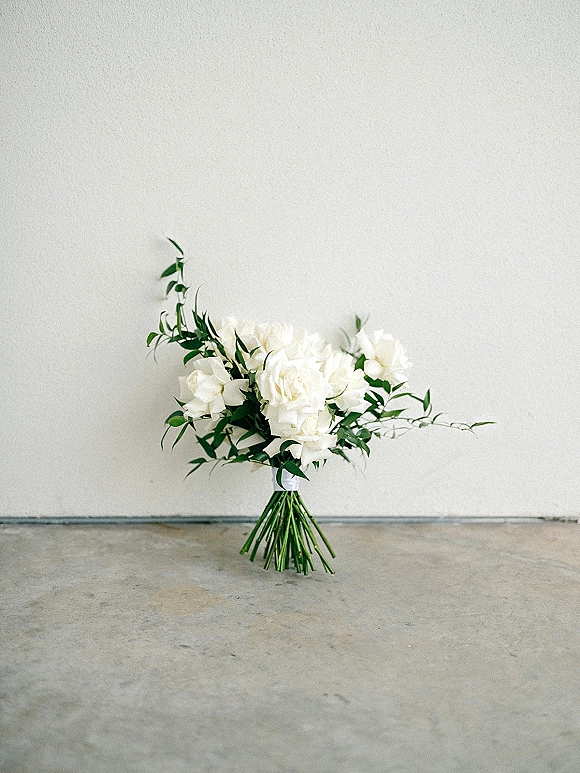 Bridal bouquet of white roses with greenery and exposed stems, arranged on a concrete floor against a white wall backdrop