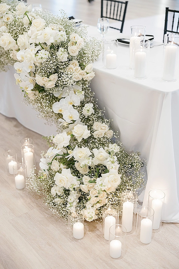 Wedding head table decor with head table floral garland of white roses, orchids, baby’s breath and greenery, plus pillar candles in glass vases