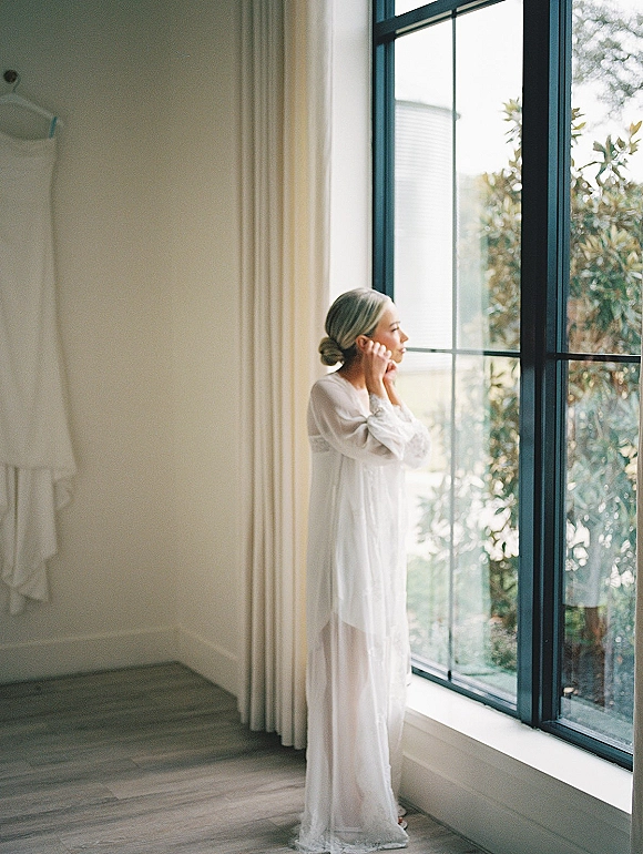 Bridal getting ready bride putting on earrings in a sheer robe by a large window, natural light on white walls with greenery outside