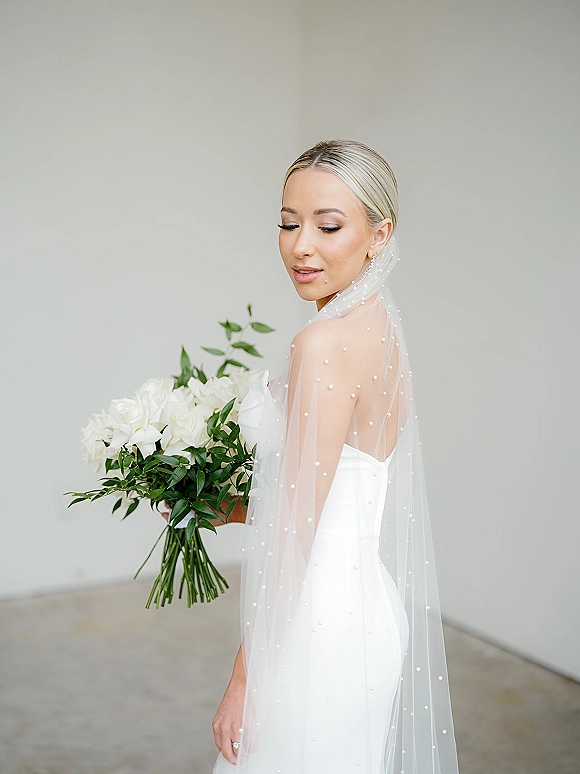 Bridal portrait of a bride in a strapless wedding dress holding a white rose and greenery bouquet, wearing a pearl veil in a studio setting