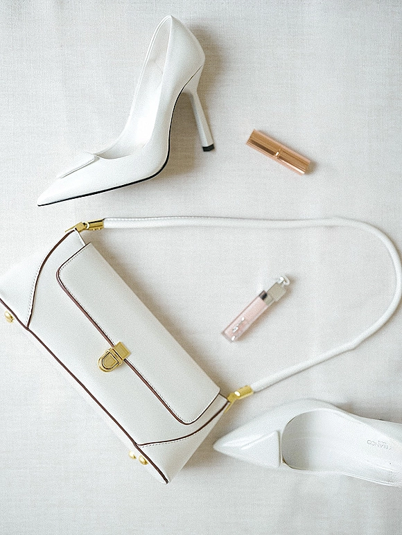Bridal accessories flat lay with white pointed toe heels, a white handbag with gold hardware, and pink lip gloss on white fabric backdrop