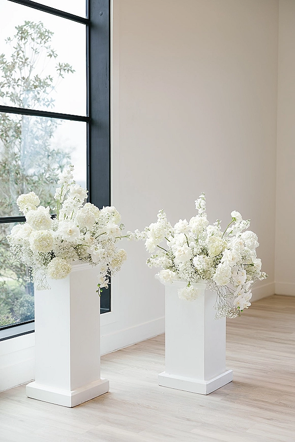 Ceremony aisle decor with wedding flower pedestals topped with white floral arrangements on stands by a large window with trees outside