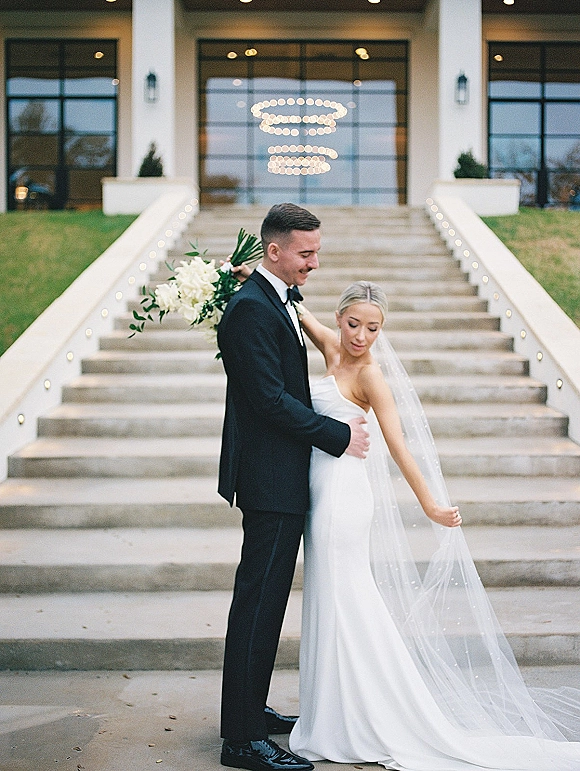 Couple portrait of bride and groom embrace on a grand staircase, bride in strapless dress with cathedral veil holding a white greenery bouquet
