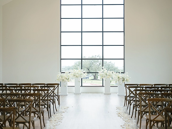 Ceremony setup with white floral arrangements on pedestals, crossback chairs, petal-strewn aisle and candle holders by a large grid window