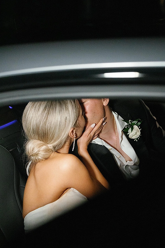Wedding kiss between bride and groom in a just married car photo, her strapless dress and his white rose boutonniere in dark backseat lighting