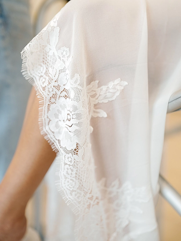 Wedding dress close-up showing lace cap sleeve appliqué on sheer tulle bodice, with a soft indoor wall and staircase railing behind