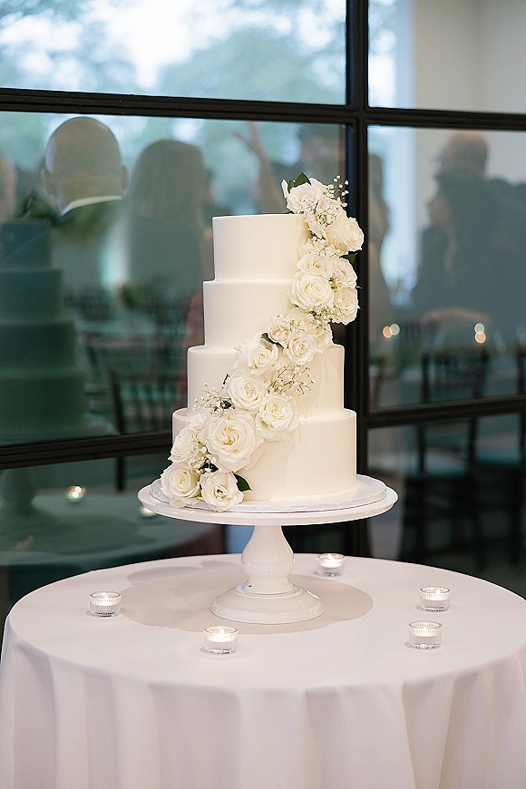 Wedding cake with smooth white buttercream and cascading white roses on a stand, surrounded by tea lights before a grid window at reception