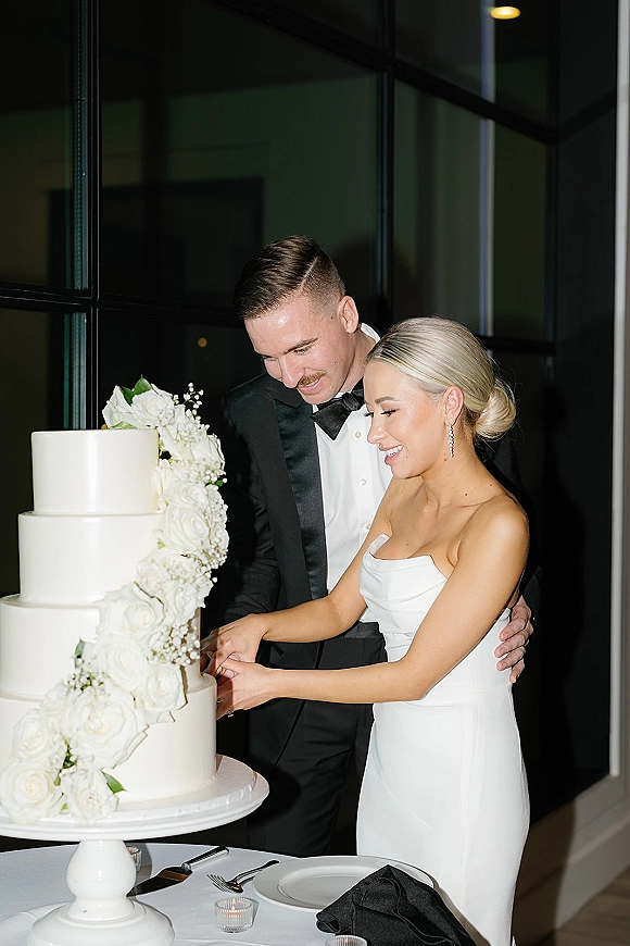 Cake cutting as bride in strapless gown and groom in tux slice a white tiered wedding cake with cascading white roses by a window wall