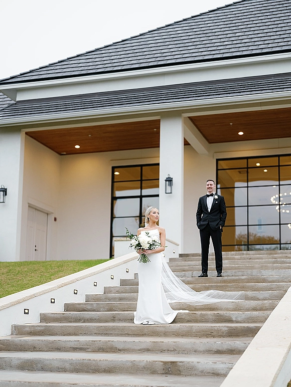 Wedding couple portrait with bride holding bouquet and groom in tuxedo on stone steps outside a modern building with grid windows under overcast sky