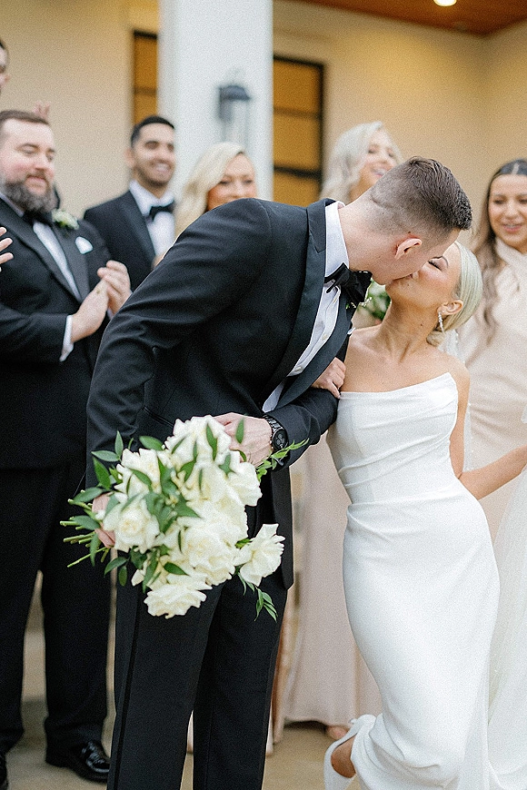Wedding kiss portrait of bride and groom kissing while walking with a white rose bouquet, wedding party cheering outside columned building