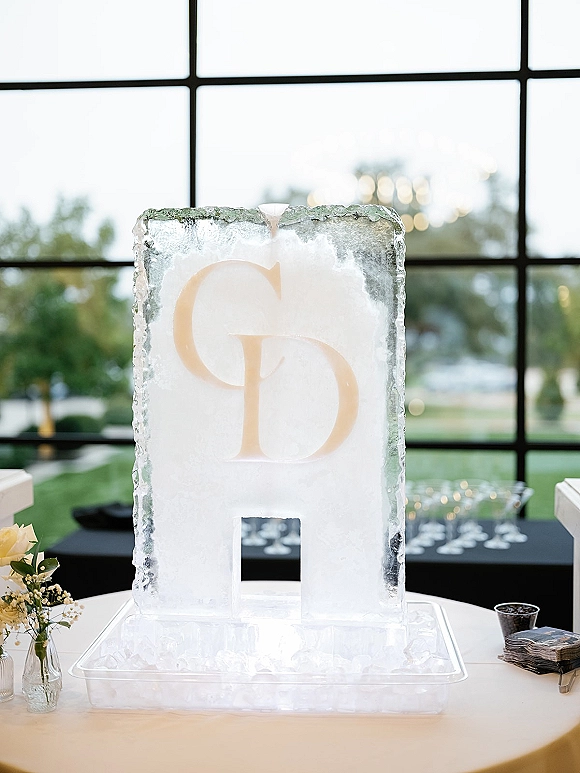 Ice sculpture wedding ice sculpture with carved monogram letters on a clear drip tray, beside white roses and napkins at a windowed bar setup