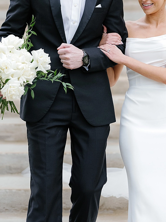 Couple portrait of bride and groom walking down stone steps, her arm linked with his, holding a white rose bouquet with greenery