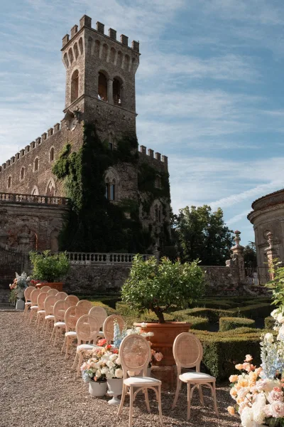 Outdoor ceremony setup featuring garden ceremony seating with cane-back chairs along a flower-lined aisle, framed by a castle and manicured hedges.