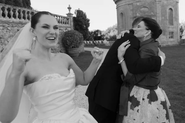 Wedding candid moment with the groom hugging mom while the bride cheers nearby, his tuxedo sharp against a stone wall and garden trees.