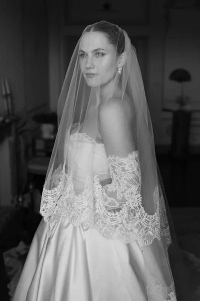 Bridal portrait of a bride with veil, her lace veil softly framing her face as she poses in a dark interior with furniture.