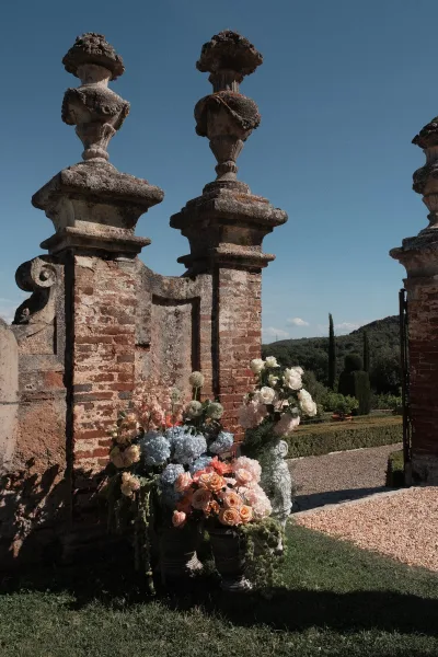 Ceremony flowers styled as garden ceremony decor with urn vases flanking a brick wall, cypress trees lining a gravel path.