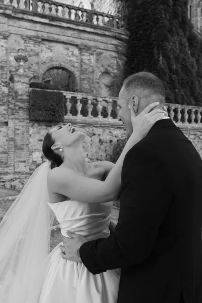 Couple portrait, a black and white wedding portrait of the bride laughing as the groom holds her waist, his black tuxedo by an ivy-draped stone wall.