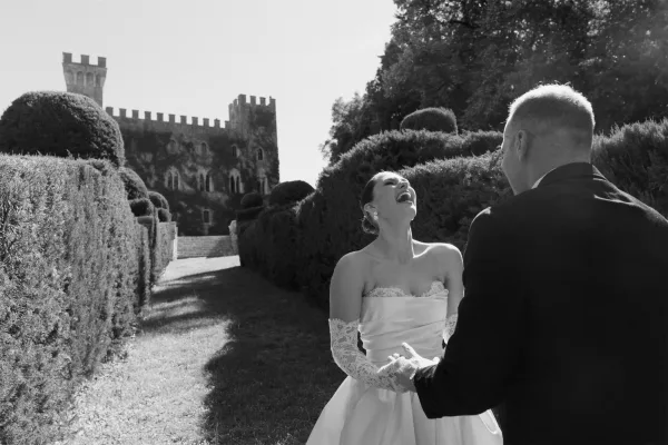 Couple portrait in a black and white wedding photo, bride wearing lace gloves beside her groom, framed by a castle and manicured hedges.