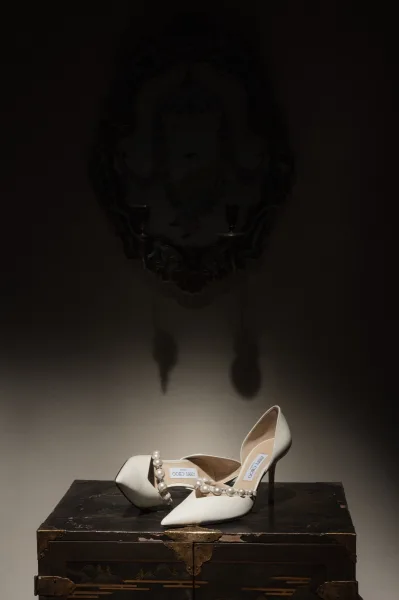 Bridal shoes, elegant pearl strap heels resting on an antique chest before an ornate mirror against a neutral wall.