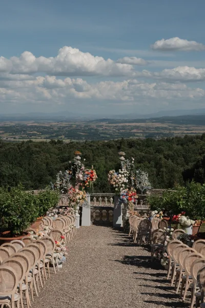 Ceremony setup showcasing refined outdoor ceremony decor with floral pillars lining a gravel aisle, framed by a stone balustrade and distant hills.