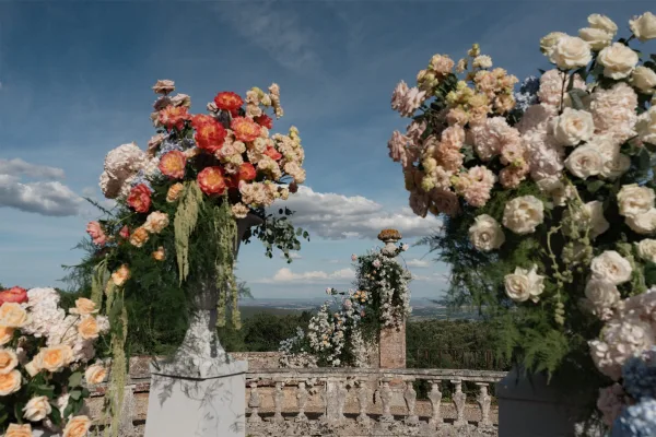 Ceremony floral decor showcasing outdoor ceremony flowers in ornate floral urns, framing a stone balustrade with blue sky and distant hills.