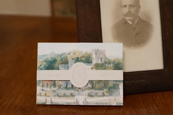 Wedding invitation featuring a watercolor wedding invitation and monogram crest, styled on a wood table beside a small framed photo.