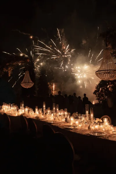 Reception fireworks erupt in an evening fireworks show above the celebration, their glow mirrored by chandeliers as smoke drifts across the night sky.