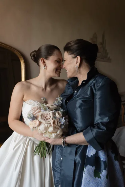 Bride and mother share a tender mother of the bride photo, hugging as the bride holds a bridal bouquet beside a mirror in a softly lit room.