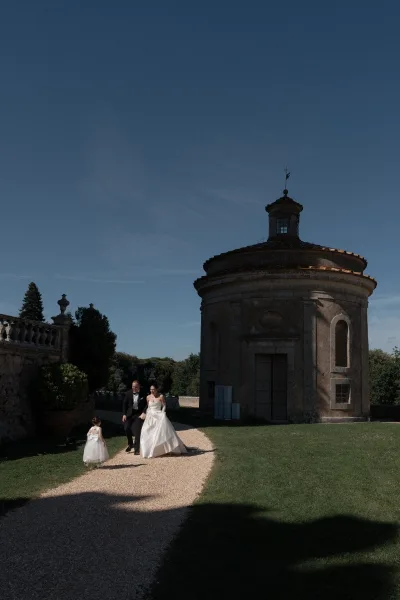 Bride and groom walking with flower girl along a gravel path, framed by a stone rotunda and balustrade amid garden trees under blue sky.