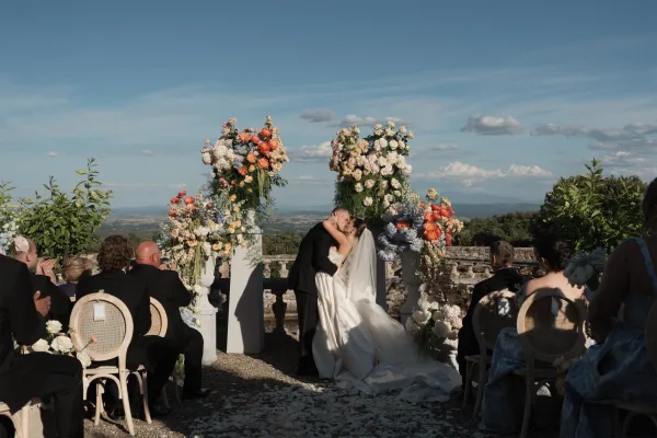 First kiss moment as the bride and groom embrace beneath vibrant floral pillars, with a blue sky and mountain landscape framing the ceremony.
