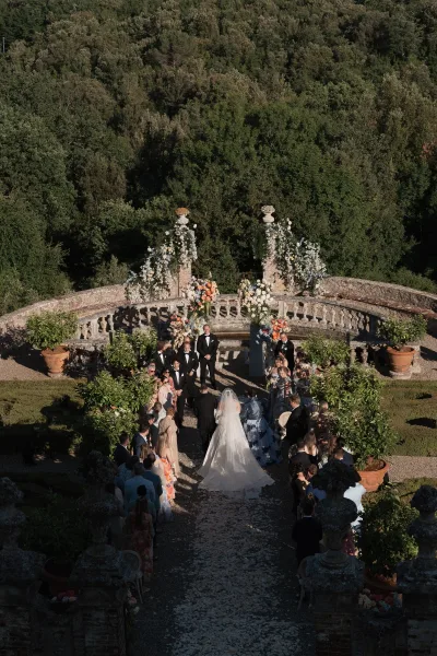 Ceremony processional with the bride walking down aisle past flower petals, guests watching on a stone terrace framed by garden trees.