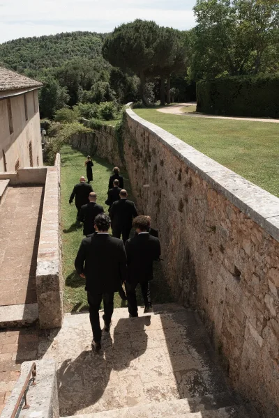 Groomsmen walking in a candid groomsmen moment down stone steps beside a weathered stone wall, wearing black suits amid a garden lawn and trees.