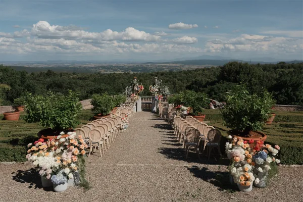 Ceremony setup with outdoor ceremony decor highlighted by floral pillars lining a gravel path, framed by a terrace balustrade and distant hills.