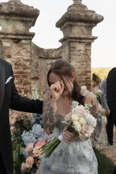Bridesmaid moment as a bridesmaid wiping tears clutches a blush rose bouquet against a rustic stone ceremony backdrop.