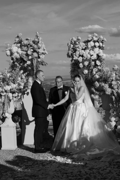Ceremony moment as the couple share wedding vows, holding hands on a stone terrace, framed by lush floral arrangements under open sky.