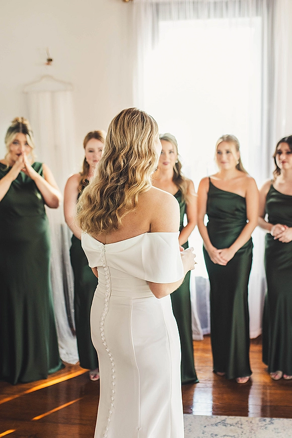 Bridesmaid reveal as bride reveals dress to bridesmaids in a bright bridal suite, off-the-shoulder button-back gown by window