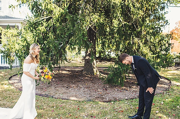 First look moment as bride reveals dress to groom, both laughing on a garden path, bouquet and veil against evergreen trees and autumn leaves