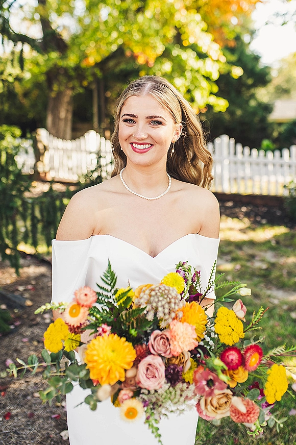Bridal portrait of bride holding bouquet in an off the shoulder wedding dress, smiling in sunlit garden by white picket fence