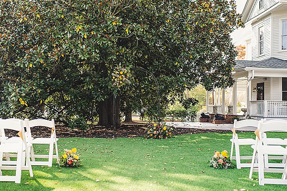 Ceremony setup with white folding chairs lining a lawn aisle toward a circular floral arch under a large tree near a sunlit porch