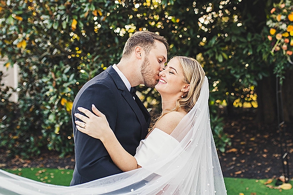 Wedding kiss portrait of bride and groom kissing, her veil flowing as she smiles in soft daylight against lush garden greenery backdrop
