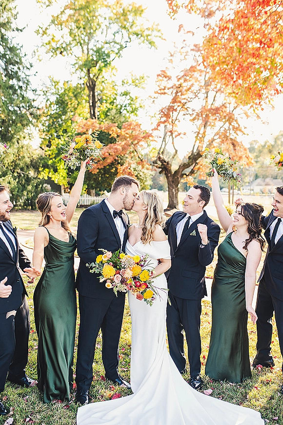 Wedding party portrait of bride and groom kissing as bridesmaids in emerald green dresses cheer with bouquets on a sunny autumn lawn